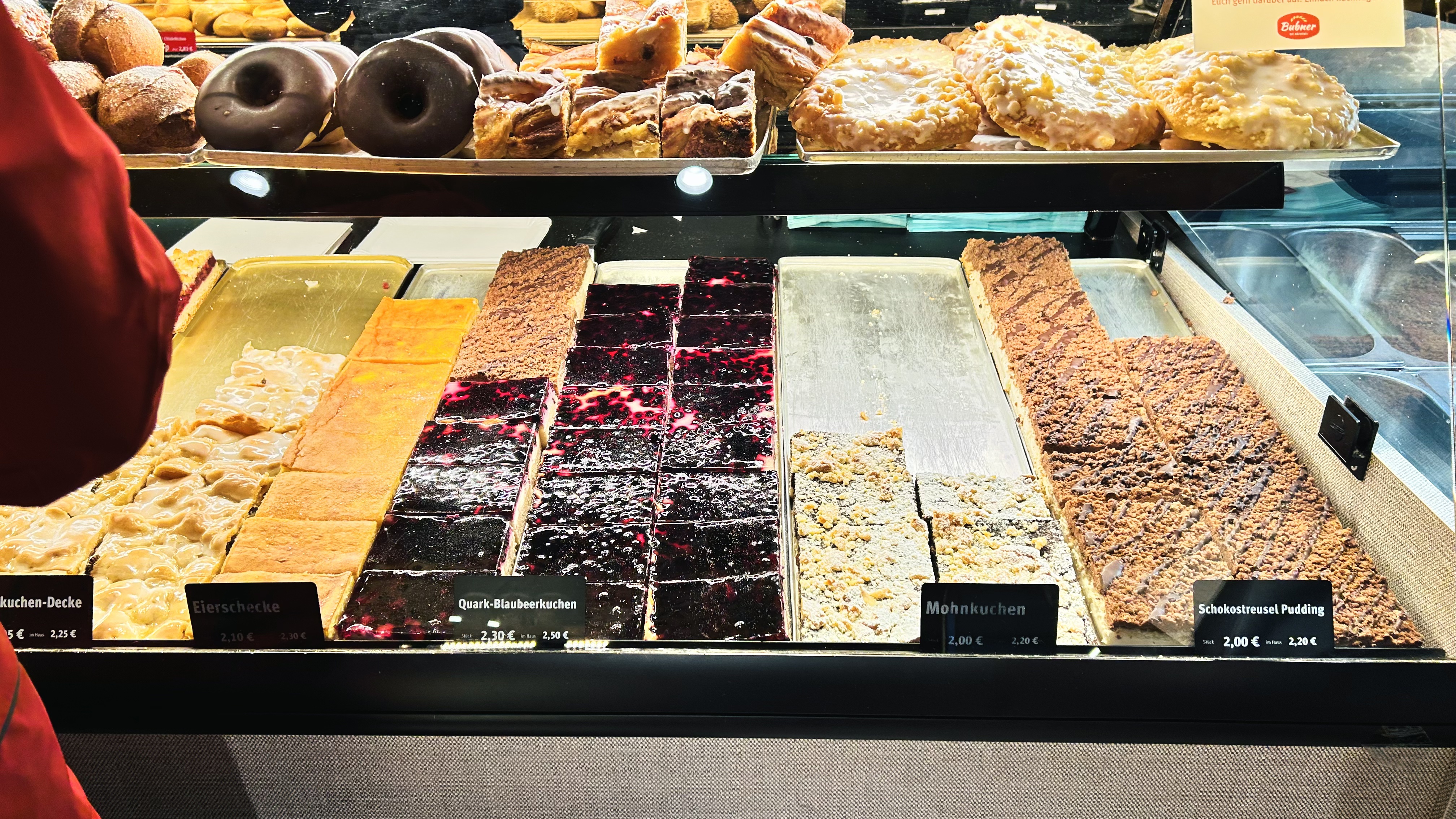 A bakery counter full of sheet cakes, including cherry crumble and poppy seed cake, with doughnuts and pastries on the shelf above.